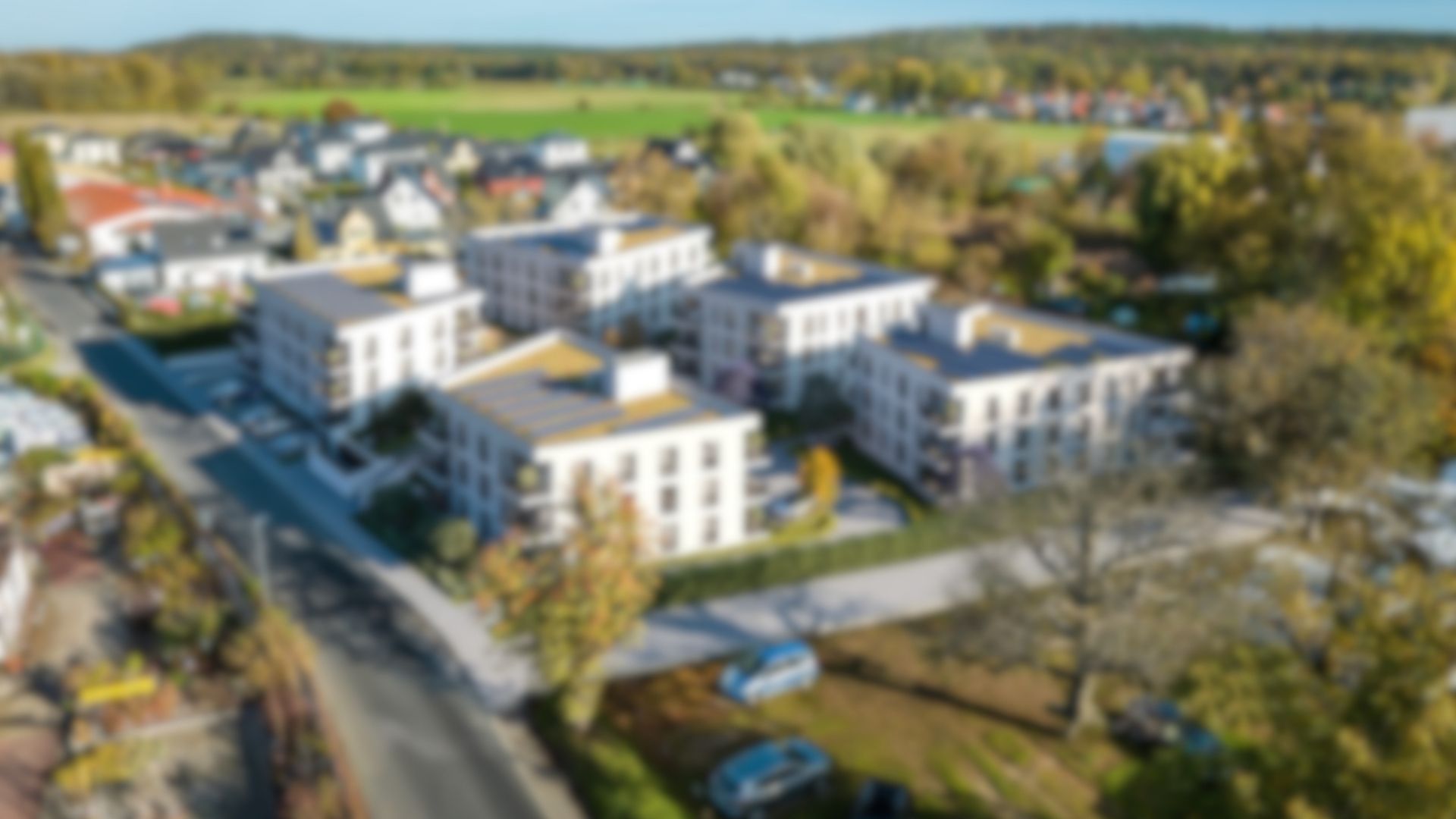 Unscharfe 3D-Architekturvisualisierung eines modernen Wohnquartiers aus der Vogelperspektive. Die Luftaufnahme zeigt ein geplantes Neubauprojekt mit mehreren hellen Mehrfamilienhäusern, Flachdächern, Balkonen und begrünten Außenanlagen, eingebettet in eine ländliche Vorstadtumgebung.